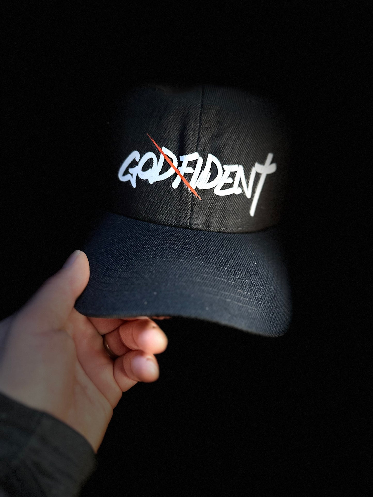 Black cap with 'GODFIDENT' text held by a hand against a dark background